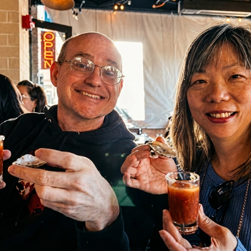 Smiling couple holding oyster shooters and oysters in a lively restaurant setting.