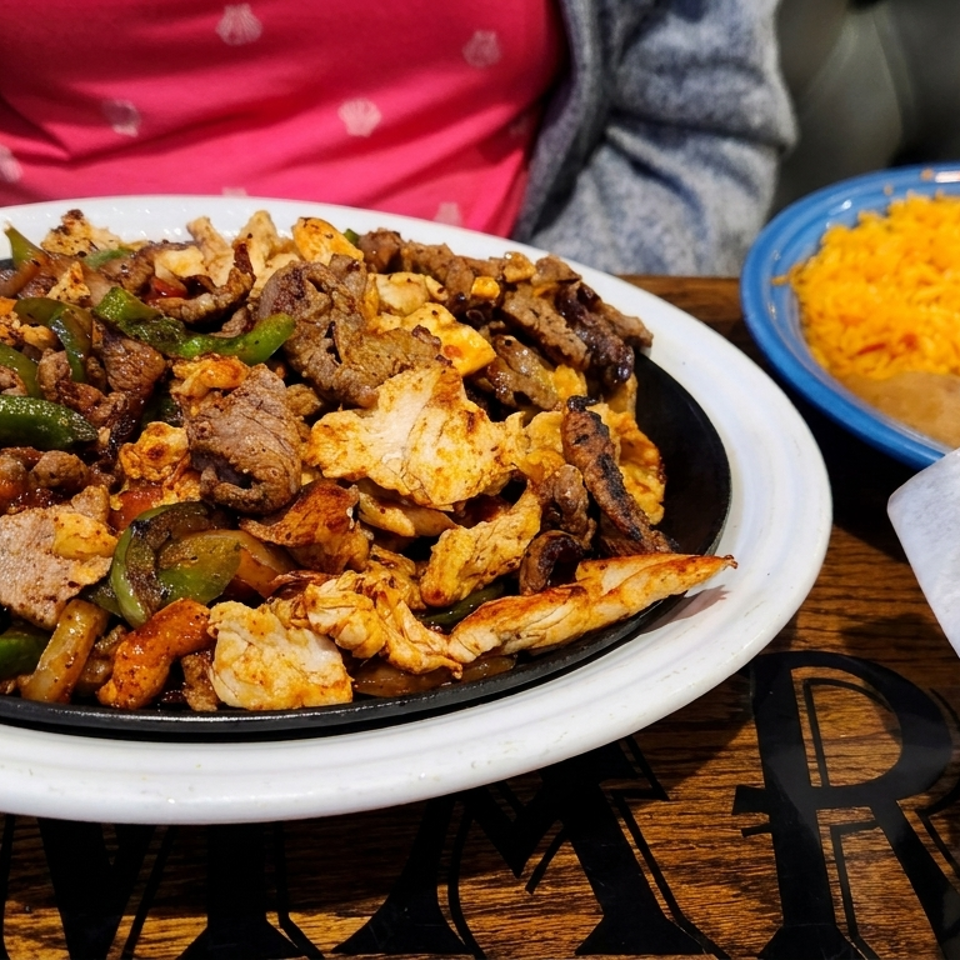 Sizzling fajitas with chicken, steak, peppers, and onions, alongside rice, beans, and tortilla chips.